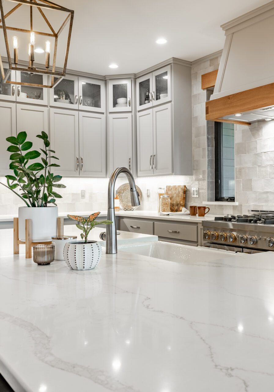 Modern kitchen with gray cabinets, stainless steel fridge, large white island with sink, built-in stove, and wooden hood. Hanging light fixture and potted plants on the countertop.