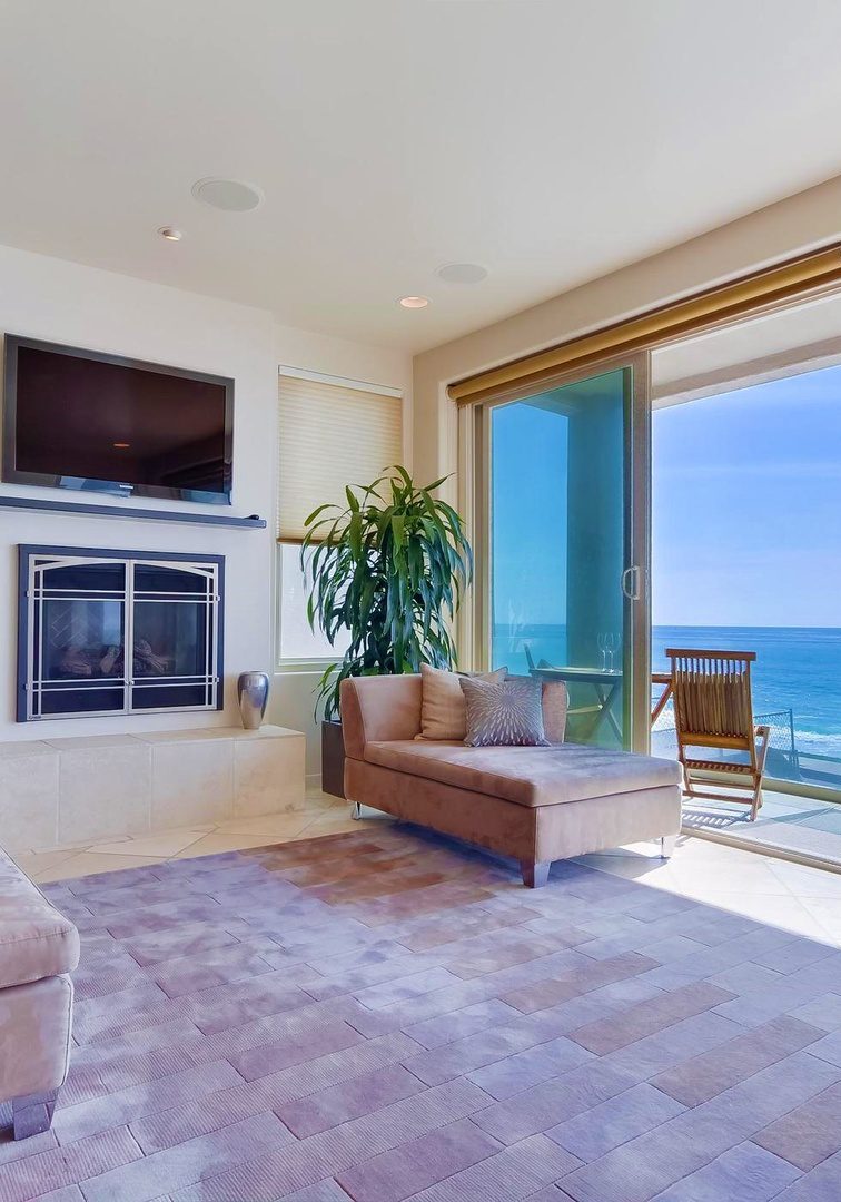 Luxurious living room with modern furniture, a wall-mounted TV above a fireplace, large windows offering an ocean view, and a small balcony with a chair.