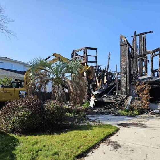 A partially collapsed, charred house with exposed wooden beams. A construction vehicle is parked nearby, and the lawn is green with a few shrubs and a palm tree.