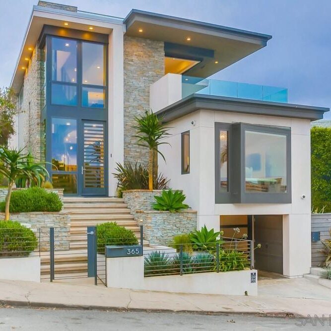 Modern multi-story house with large windows, stone and stucco exterior, landscaping, and a small front gate, situated on a sloped street.